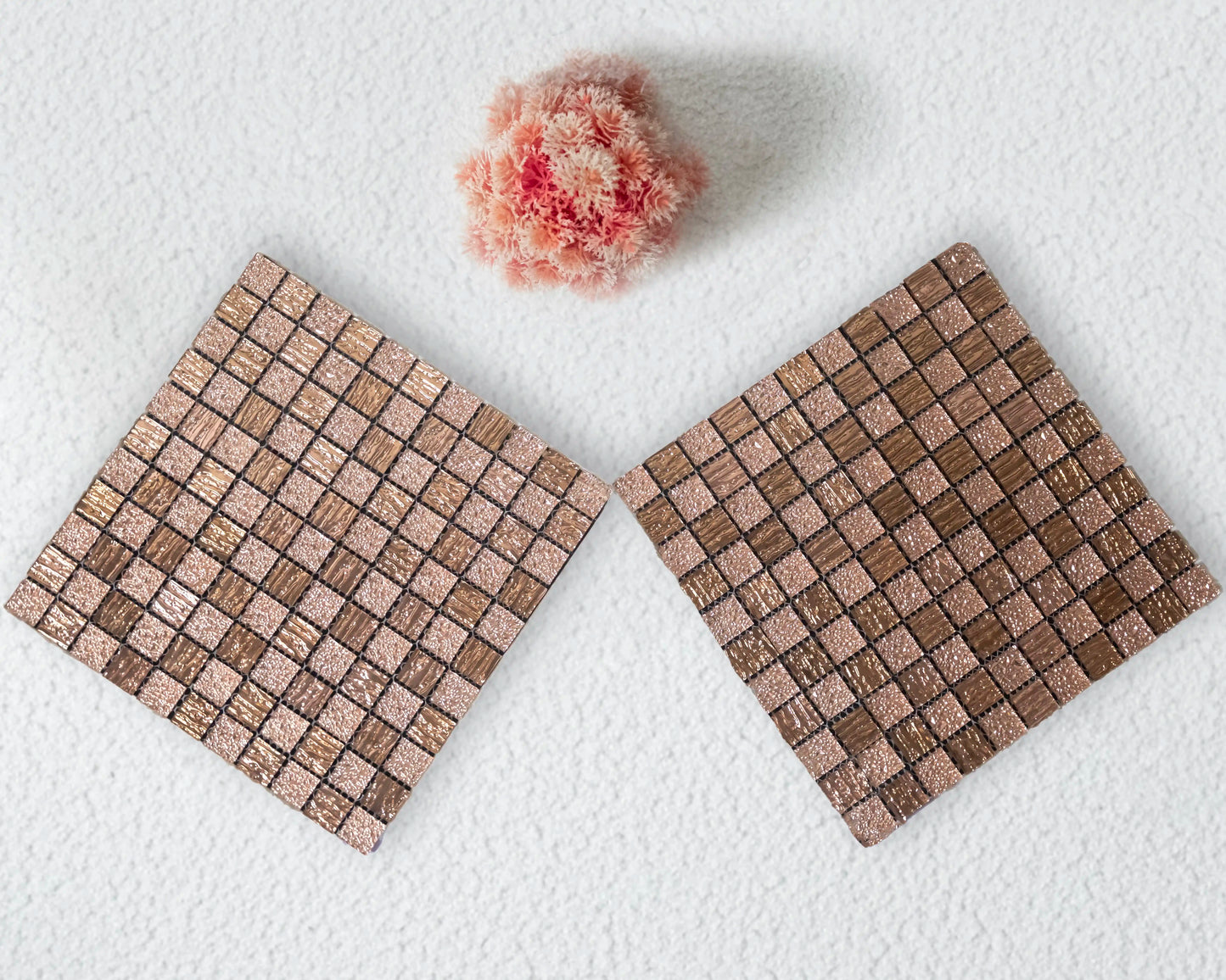 Two square woven coasters on a white surface with a small pink flower.