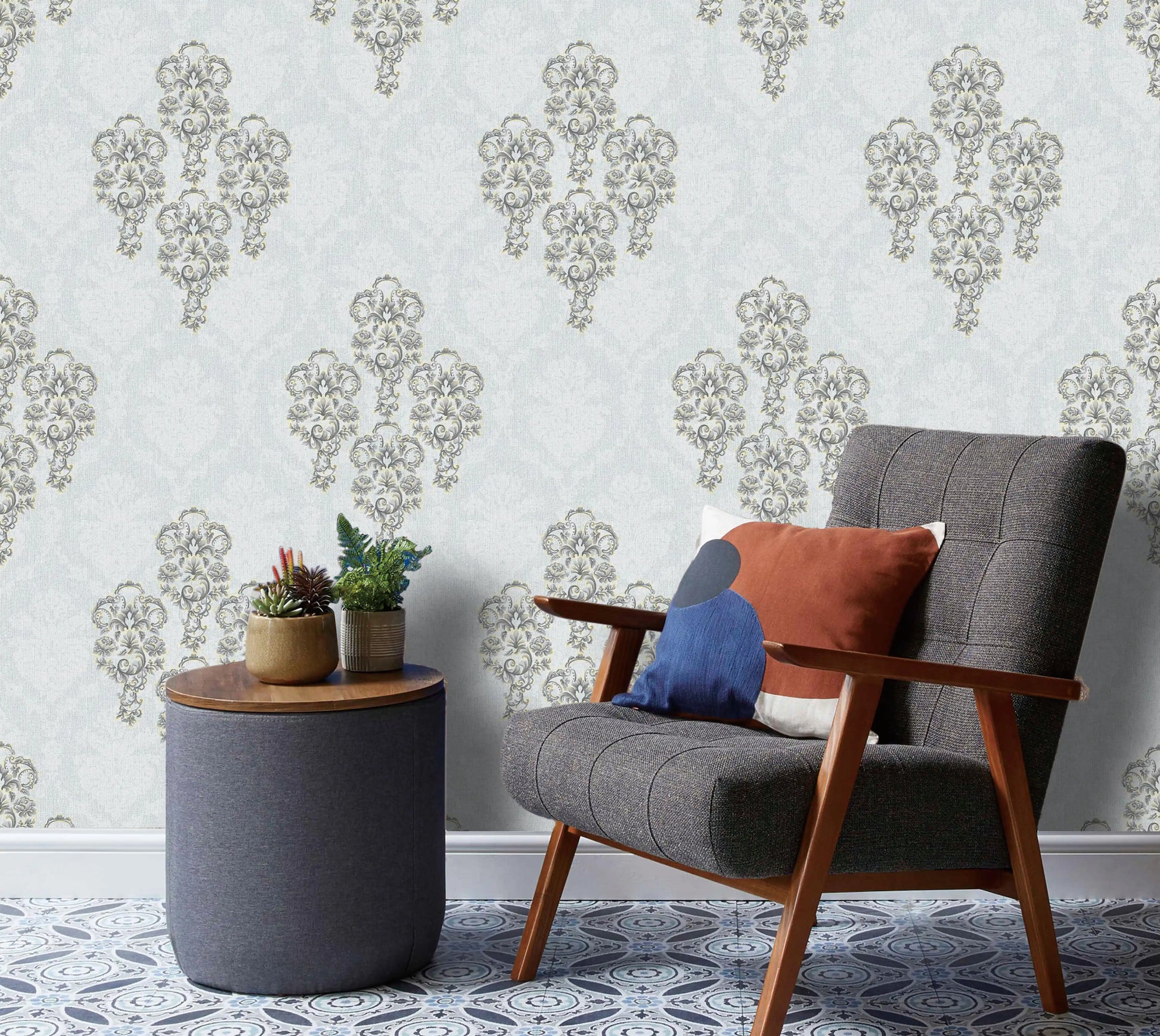 Gray armchair with colorful cushions next to a small round table with plants against a decorative wallpaper.