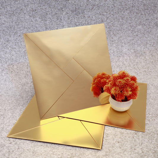 Several golden self-adhesive acrylic mirror mosaic tiles are displayed against a grey background, with one tile standing vertically next to a small round decoration featuring orange flowers in a white pot.