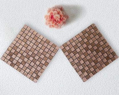 Two square woven coasters on a white surface with a small pink flower.